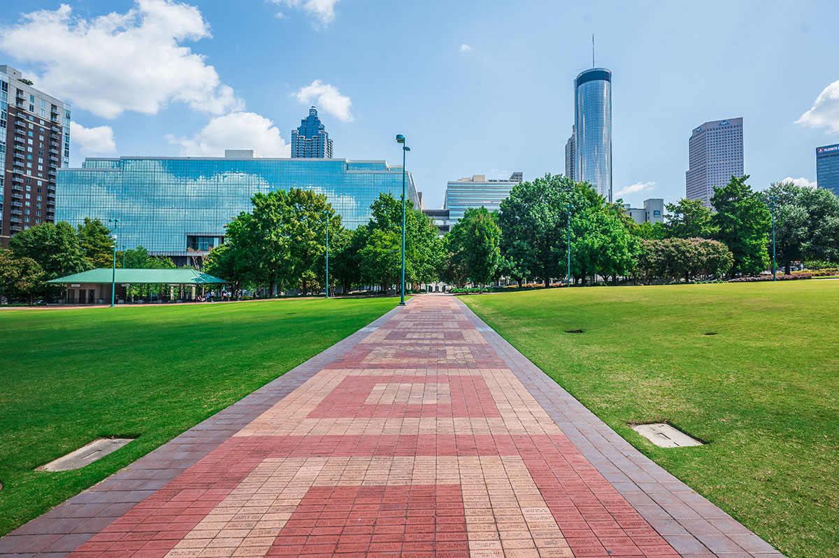 A brick pathway runs through a green park toward city buildings and skyscrapers under a bright blue sky with scattered clouds.