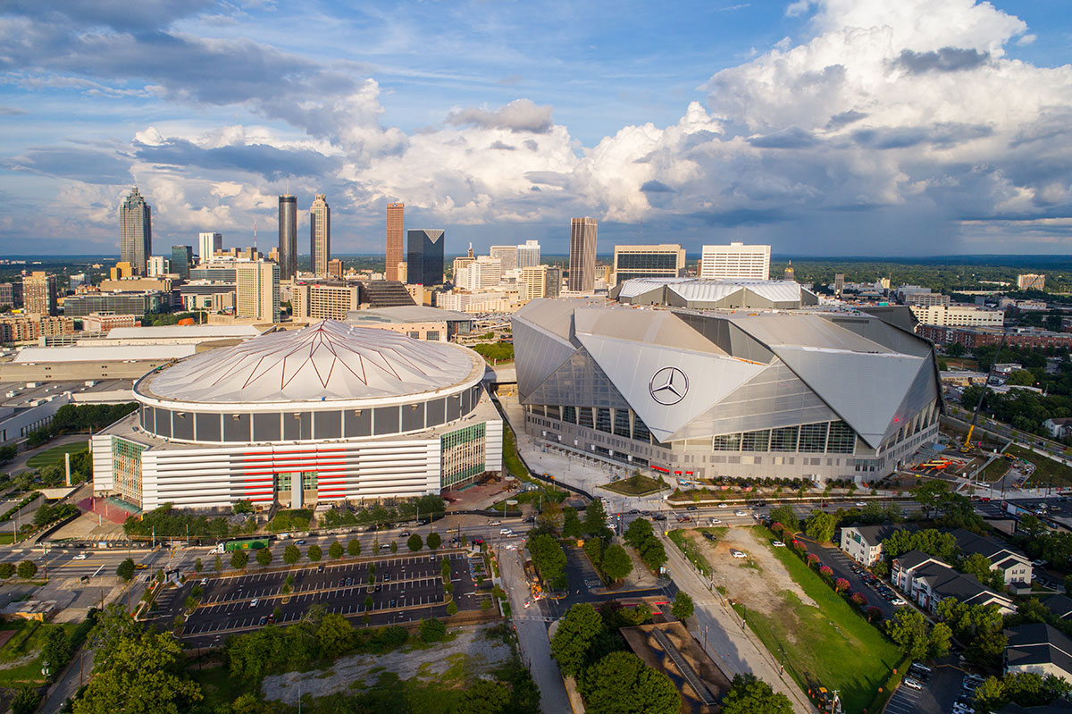 Aerial view of two large stadiums in a city: one is circular with a striped roof, the other has a modern angular design with a Mercedes-Benz logo. Downtown skyscrapers and scattered clouds are in the background.