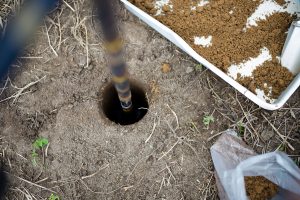 A metal pole is inserted into a round hole in the soil. Nearby, there is a tray filled with dirt and a plastic bag on the ground with dry grass and small green plants scattered around.