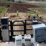 Workers in safety vests plant seedlings in a muddy field, marked by stakes and cones. In the foreground, bags and containers of agricultural supplies sit on a metal platform.