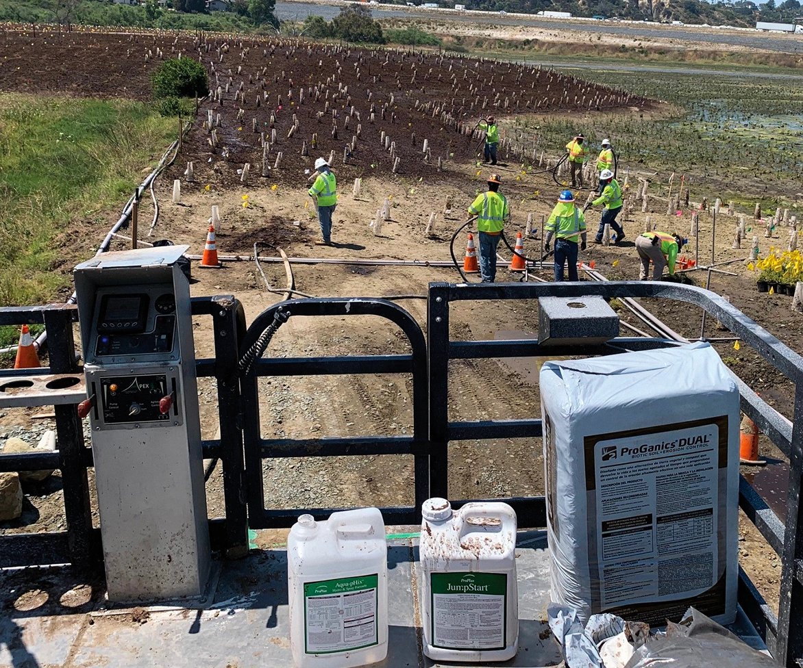 Workers in safety vests plant seedlings in a muddy field, marked by stakes and cones. In the foreground, bags and containers of agricultural supplies sit on a metal platform.