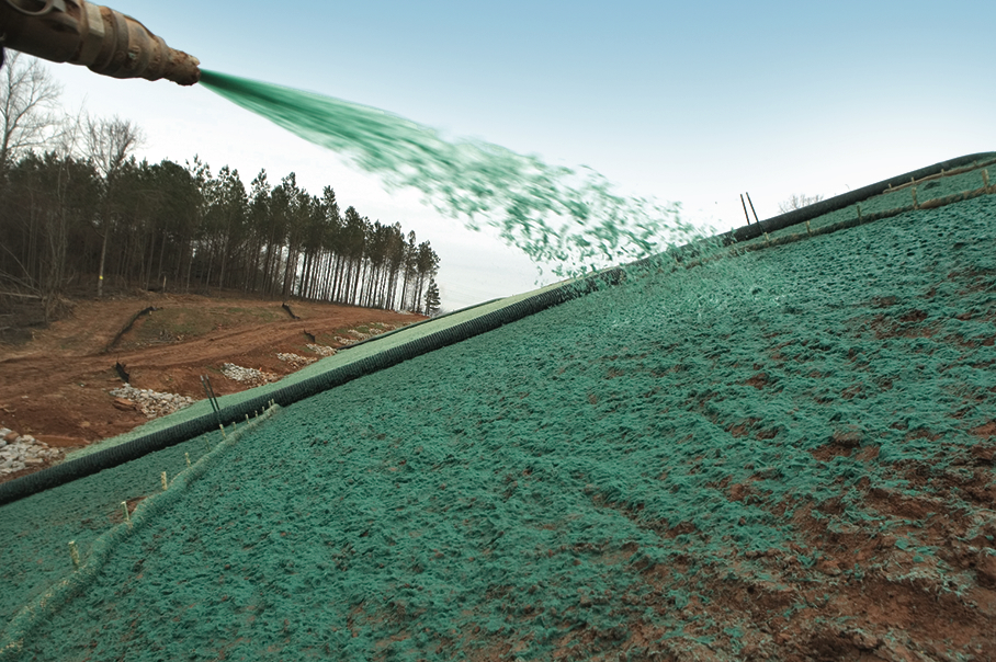 A hose sprays green hydroseed mixture onto a sloped, bare dirt embankment for erosion control, with a line of trees and a dirt road in the background.