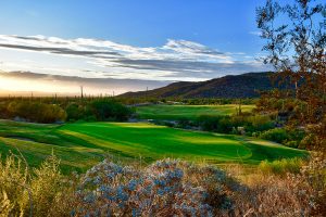 A green golf course surrounded by desert vegetation, cacti, and hills under a partly cloudy sky at sunset.