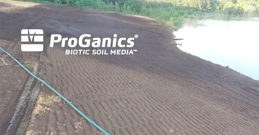 A riverbank covered with freshly spread soil media, a green hose on the ground, and trees in the background. Overlaid text reads ProGanics BIOTIC SOIL MEDIA..