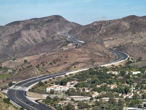 A winding freeway cuts through dry, hilly terrain with sparse vegetation, power lines overhead, and a residential area with white-roofed houses in the foreground.