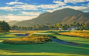 A scenic golf course with sand bunkers, green fairways, and distant houses, set against a backdrop of large, sunlit mountains and a partly cloudy blue sky.