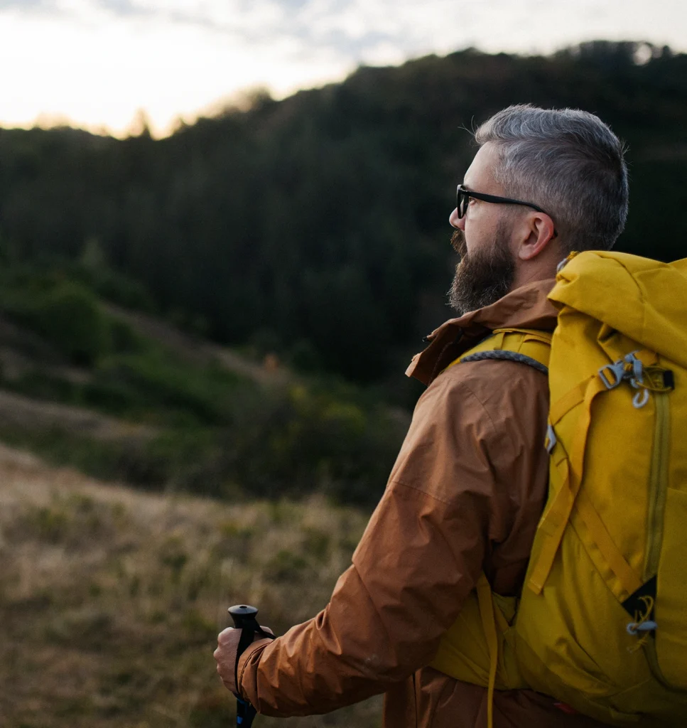 A person with gray hair and a beard, wearing glasses, a brown jacket, and a yellow backpack, stands outdoors with hiking poles, looking at distant green hills under a cloudy sky.