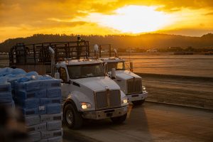 Two white trucks drive on a dusty construction site at sunset, with stacked materials in the foreground and a worker visible on a truck. The sky is bright orange and silhouetted hills are in the background.