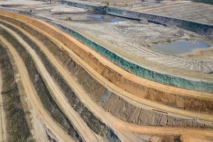 Aerial view of a large open-pit mine with terraced layers of earth, construction vehicles, exposed soil, and a few water puddles. The scene shows excavation and earth-moving activities.