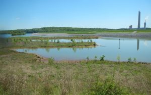 A calm reservoir with small islands and sparse vegetation in the foreground, bordered by a dam and distant smokestacks under a clear blue sky.