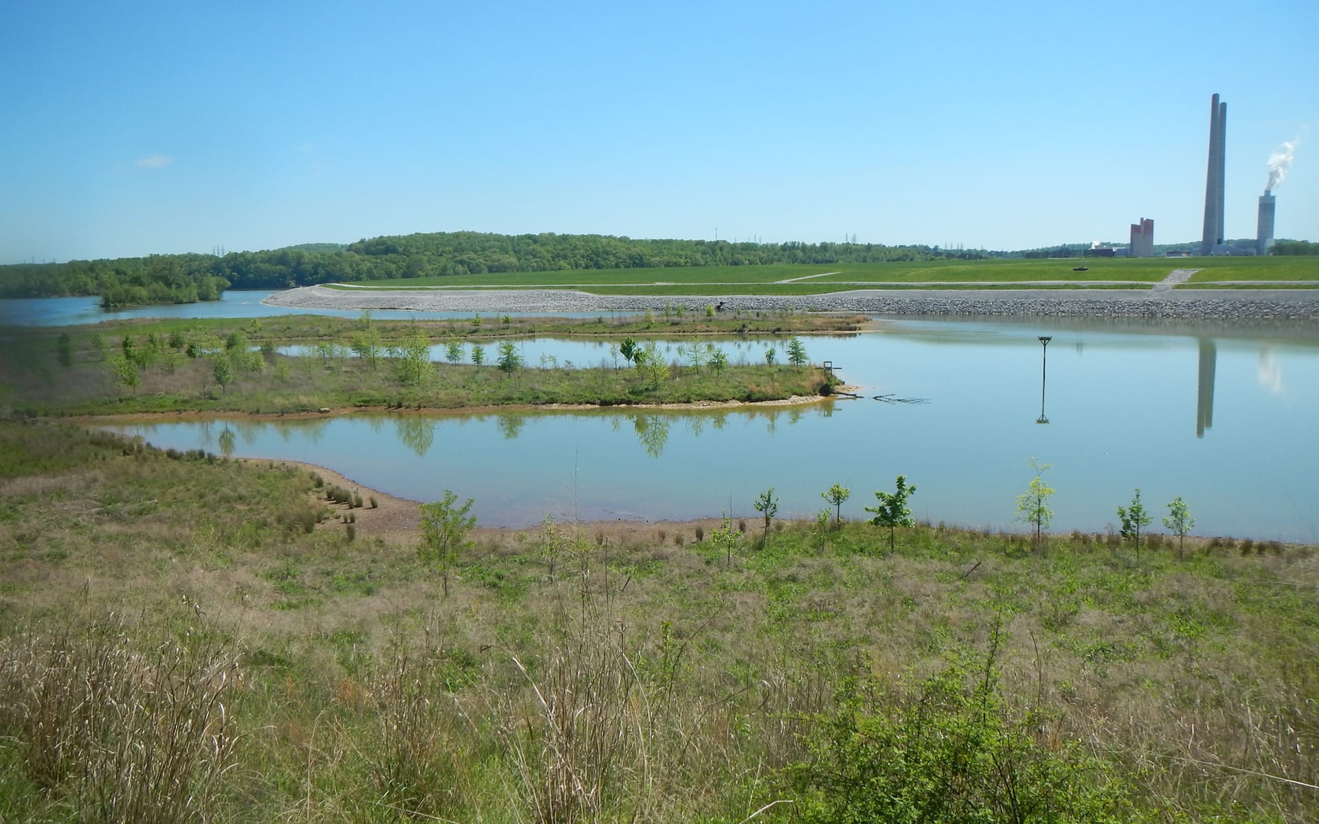 A calm reservoir with small islands and sparse vegetation in the foreground, bordered by a dam and distant smokestacks under a clear blue sky.