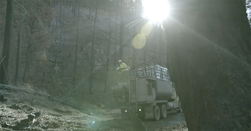 A truck with workers drives through a forested, sunlit area with bare trees. Sunlight creates lens flares, and the terrain looks rugged and burned.