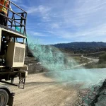 Worker on a platform operating a machine that sprays bright green liquid onto a dirt road in a mountainous landscape under a clear sky.