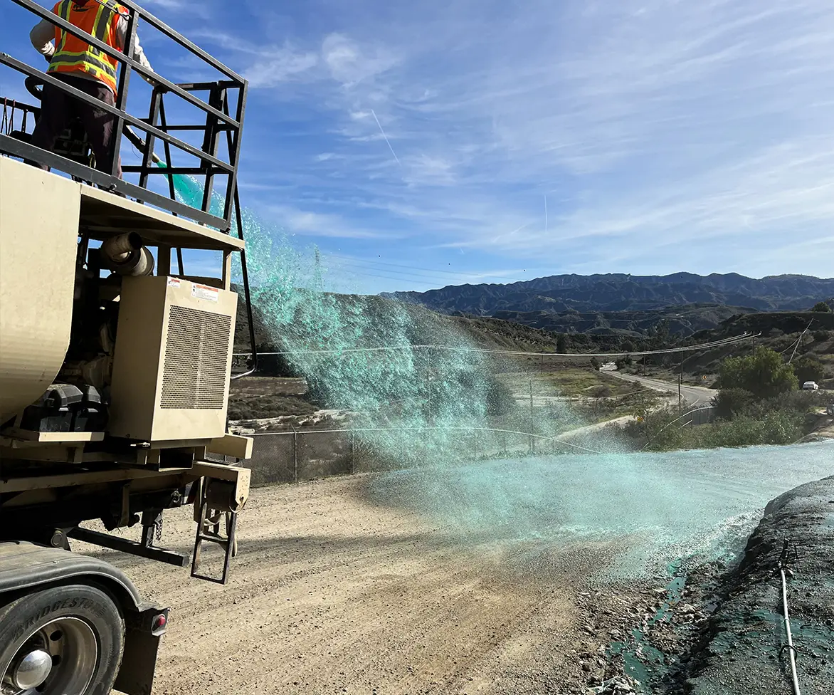 Worker on a platform operating a machine that sprays bright green liquid onto a dirt road in a mountainous landscape under a clear sky.