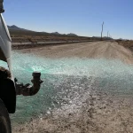 Truck nozzle spraying green liquid across a dusty rural road with power lines and hills in the distance.