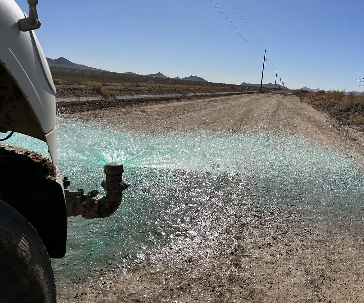 Truck nozzle spraying green liquid across a dusty rural road with power lines and hills in the distance.