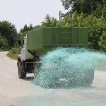 Rear view of a green tank truck spraying green liquid across a paved rural road bordered by trees.