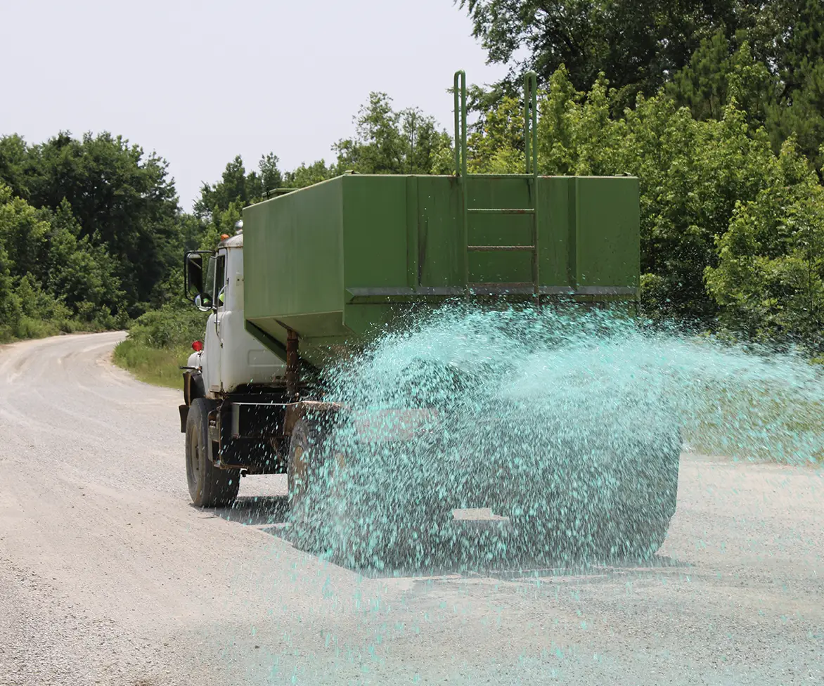 Rear view of a green tank truck spraying green liquid across a paved rural road bordered by trees.