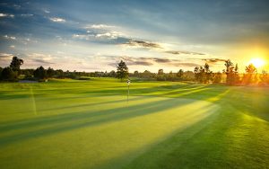Golf course at sunset with long shadows stretching across the green, flagstick in the center, trees and blue sky with clouds in the background, and the sun low on the horizon.