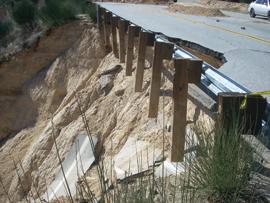 A damaged road with a guardrail is collapsing down a steep, sandy slope due to erosion or a landslide, exposing support posts and loose dirt.