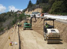 Construction workers operate heavy machinery, including a roller and excavator, while working on a dirt road cut into a hillside, surrounded by trees and construction materials.