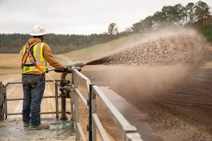 A construction worker in safety gear sprays soil or mulch from a hose onto a large open area, standing on a raised platform with grassy hills and trees in the background.