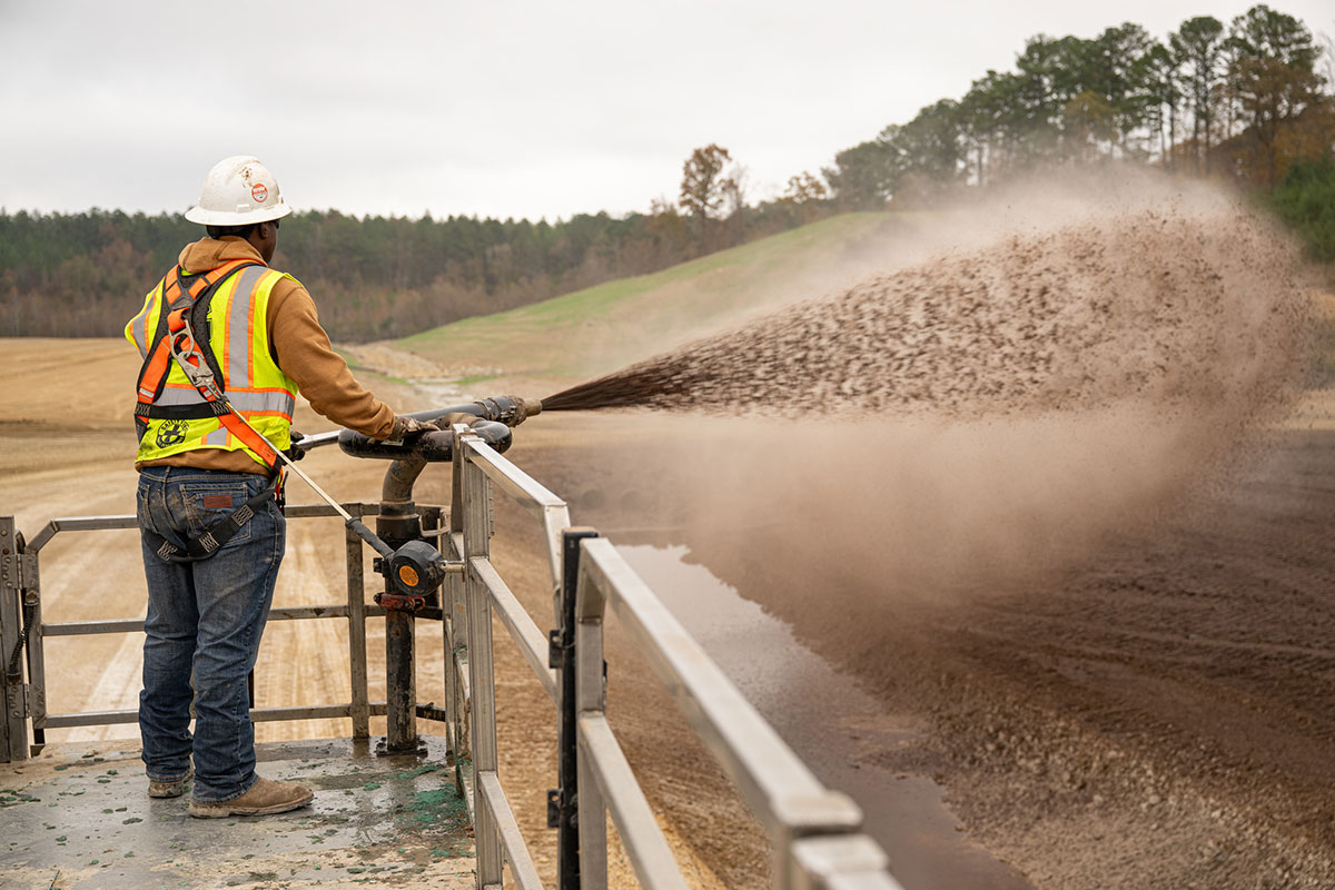 A construction worker in safety gear sprays soil or mulch from a hose onto a large open area, standing on a raised platform with grassy hills and trees in the background.