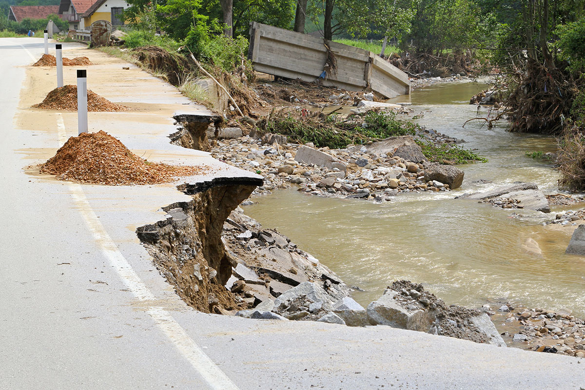 A damaged road with large sections washed away, piles of gravel, and a muddy river flowing nearby. Broken concrete barriers and debris are visible in the water and along the bank.