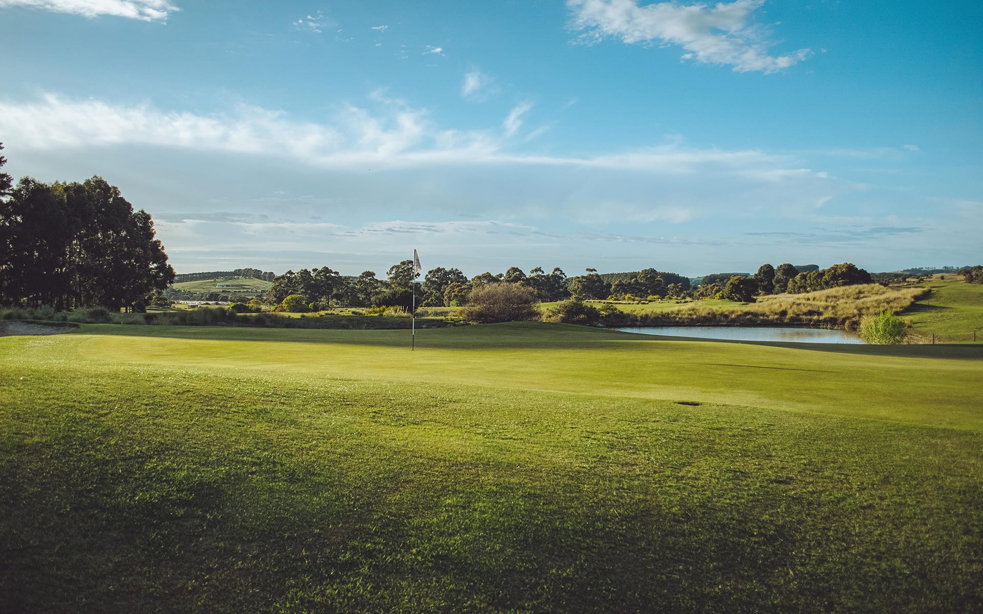 A golf course with a flag on the green, surrounded by trees, a pond, and open blue sky with scattered clouds.