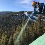 Two workers on a lift spray green hydroseed onto a slope, with a forested, snow-speckled mountain landscape in the background under a clear blue sky.
