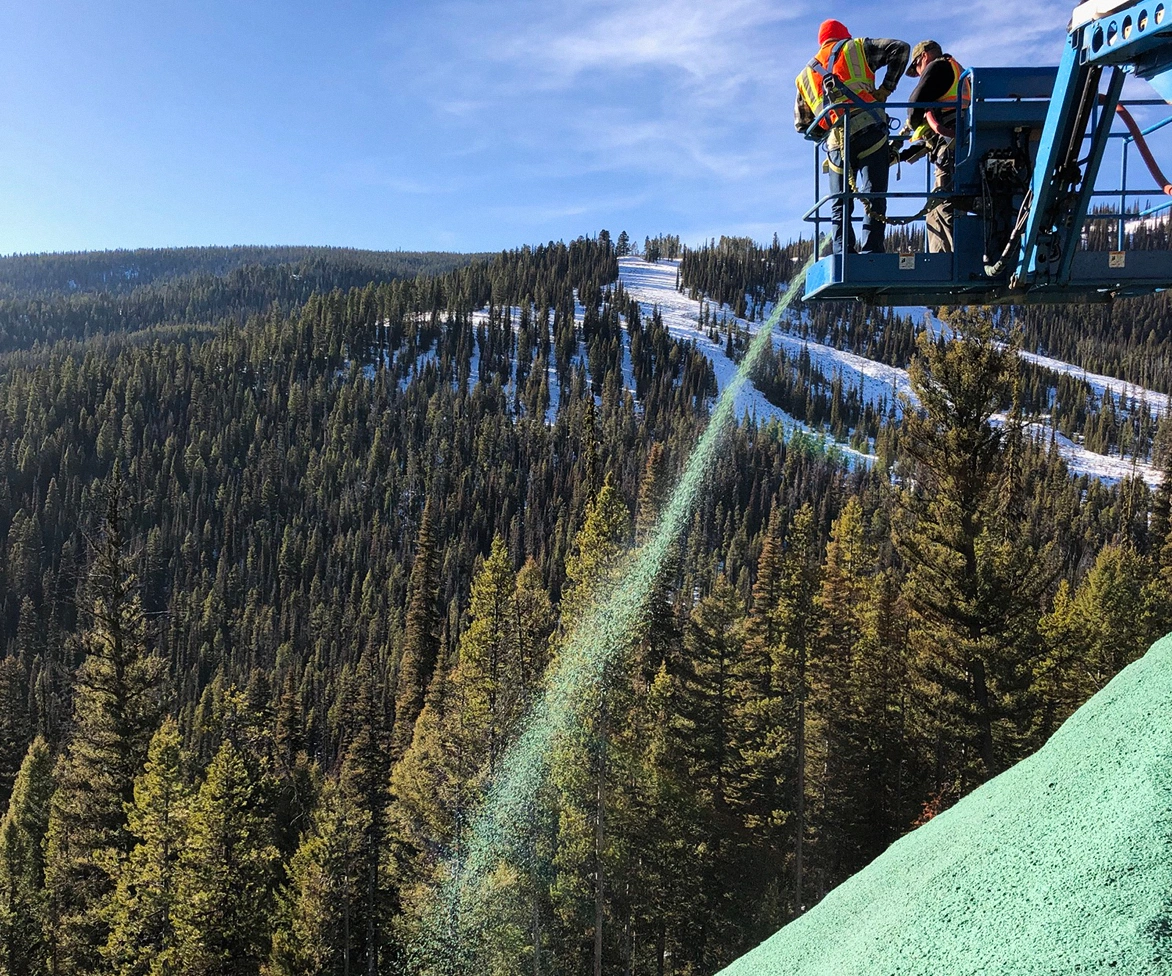 Two workers on a lift spray green hydroseed onto a slope, with a forested, snow-speckled mountain landscape in the background under a clear blue sky.