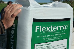 A hand holds a large white bag labeled Flexterra Flexible Growth Medium, used for erosion control on steep slopes and critical sites.