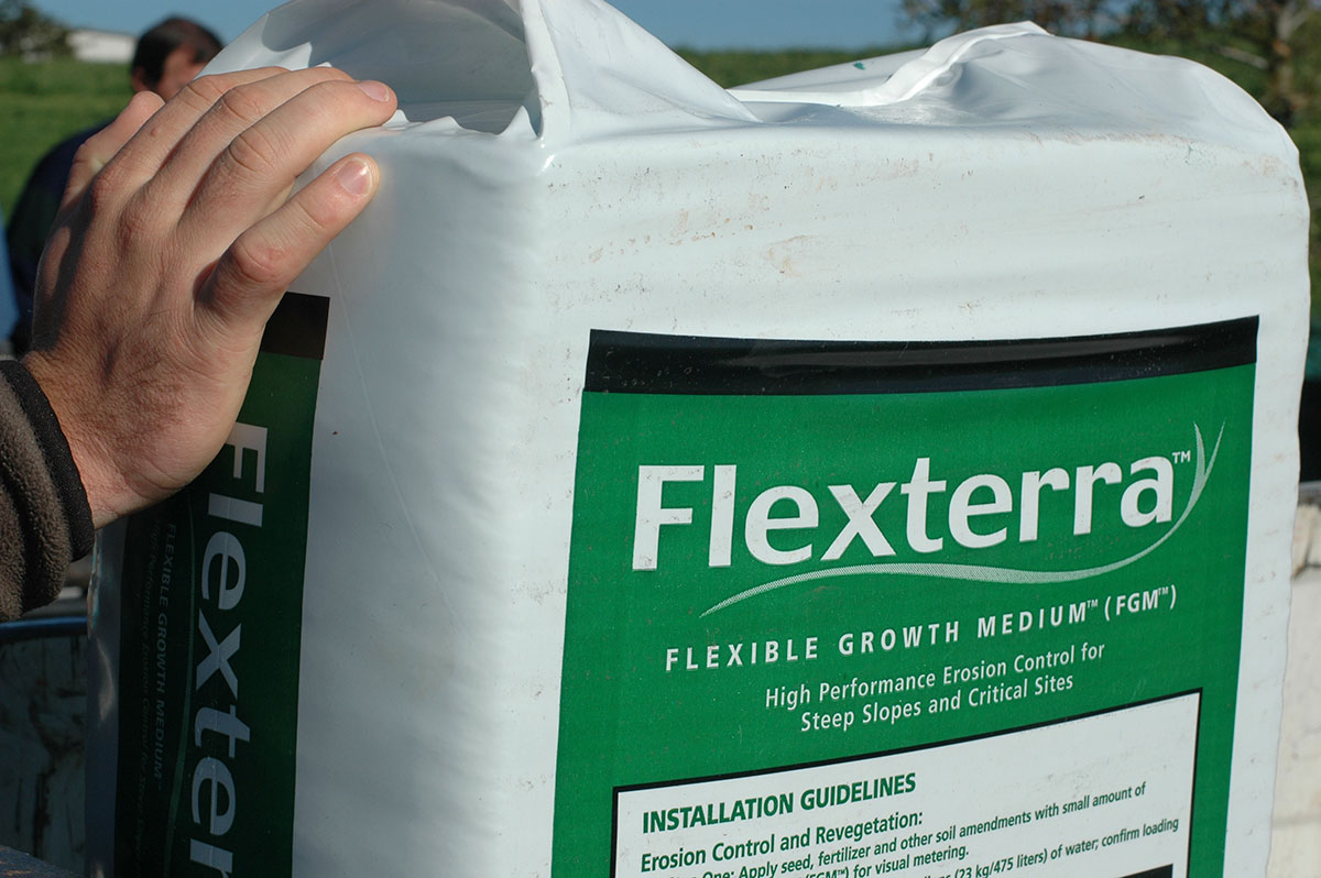 A hand holds a large white bag labeled Flexterra Flexible Growth Medium, used for erosion control on steep slopes and critical sites.