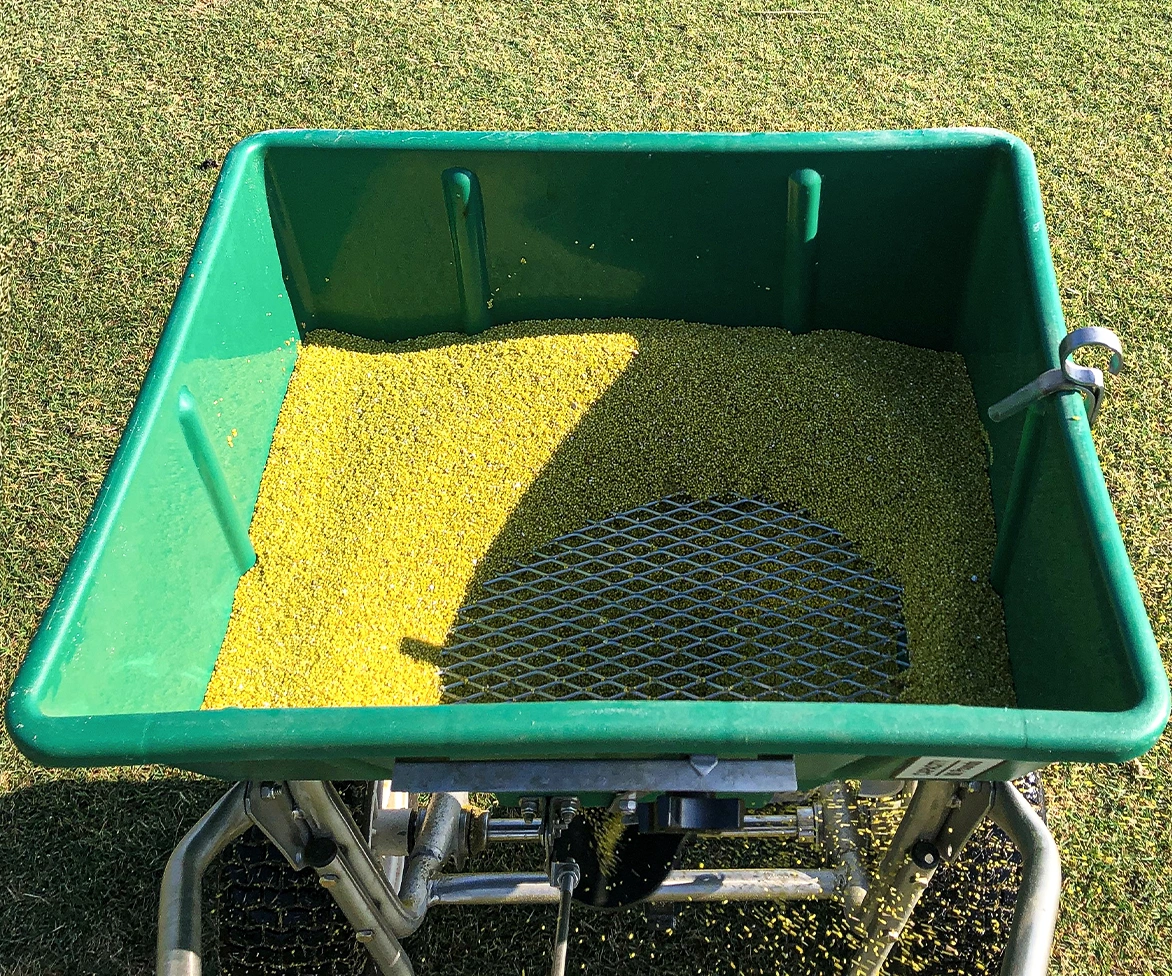 A green spreader filled with yellow granular fertilizer stands on grass, with some granules visible beneath a metal mesh inside the spreader.