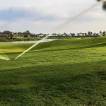 Green golf course with sprinklers watering the grass, houses and palm trees in the background, and a pond reflecting the sky on a partly cloudy day.