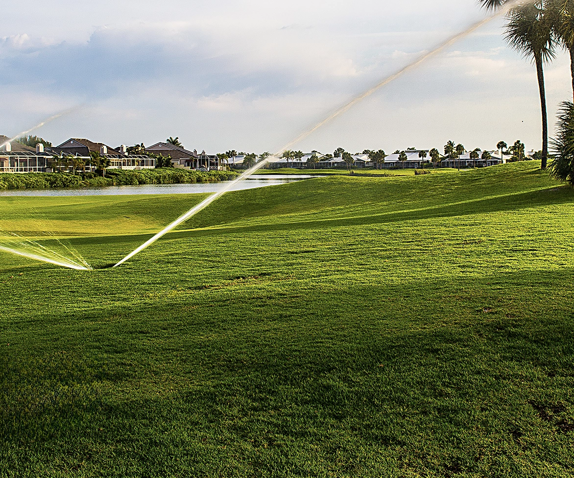 Green golf course with sprinklers watering the grass, houses and palm trees in the background, and a pond reflecting the sky on a partly cloudy day.