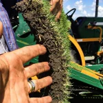 A close-up of hands holding a piece of grass turf with soil, with farming equipment visible in the background on a sunny day.