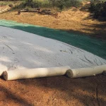 Large rolls of erosion control blanket partially unrolled over a sloped embankment beside a paved road, with a strip of green hydroseeded area along the roadside.