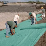 Workers installing a large green erosion control blanket across a graded slope, securing the material to the soil during landscaping or site stabilization.