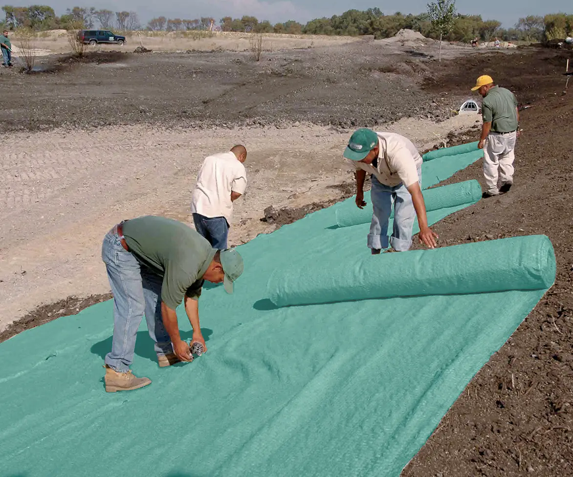 Workers installing a large green erosion control blanket across a graded slope, securing the material to the soil during landscaping or site stabilization.