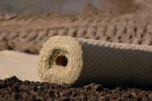 A close-up of a rolled-up erosion control mat resting on soil, with another similar mat blurred in the background. The mat appears woven, possibly made of natural fibers.