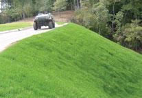 A grassy hill slopes beside a road, with a black off-road vehicle parked on the pavement near a wooded area.