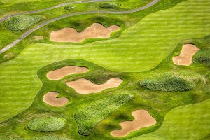 Aerial view of a golf course showing bright green grass, sand bunkers, patches of rough, and a winding gray cart path. The course appears well-maintained with striped mowing patterns.