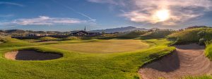 A scenic golf course with sand bunkers, green grass, rolling hills, and a bright sun setting near distant mountains under a blue sky with wispy clouds.