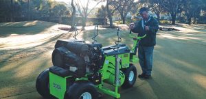 A man stands on grass operating a large green turf maintenance machine on a golf course, surrounded by trees in the early morning light.