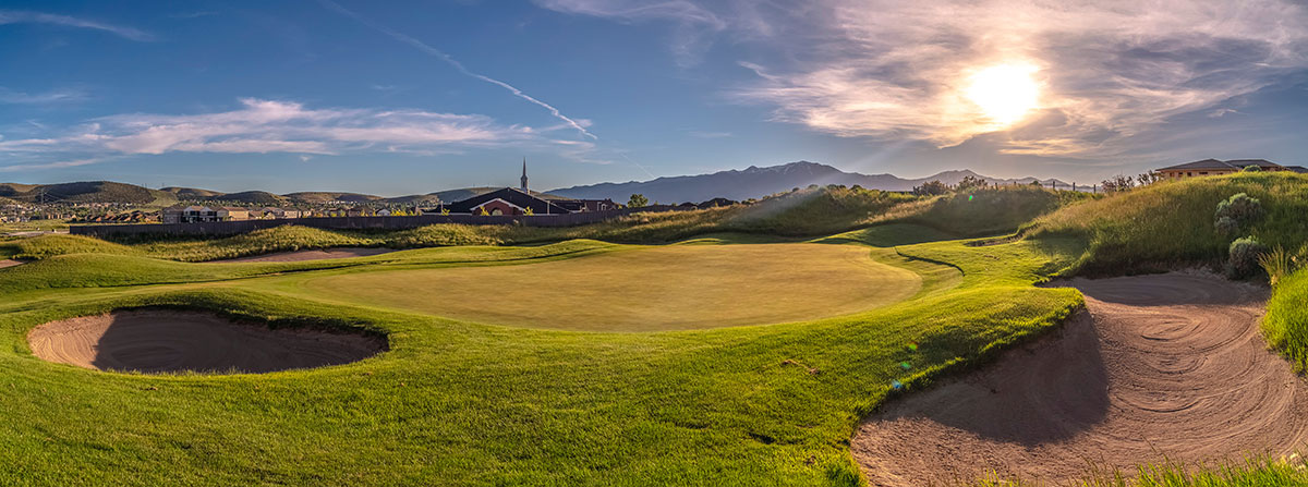 A scenic golf course with sand bunkers, green grass, rolling hills, and a bright sun setting near distant mountains under a blue sky with wispy clouds.