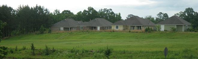 A row of single-story houses with sloped roofs is seen behind a grassy field and trees under a cloudy sky.