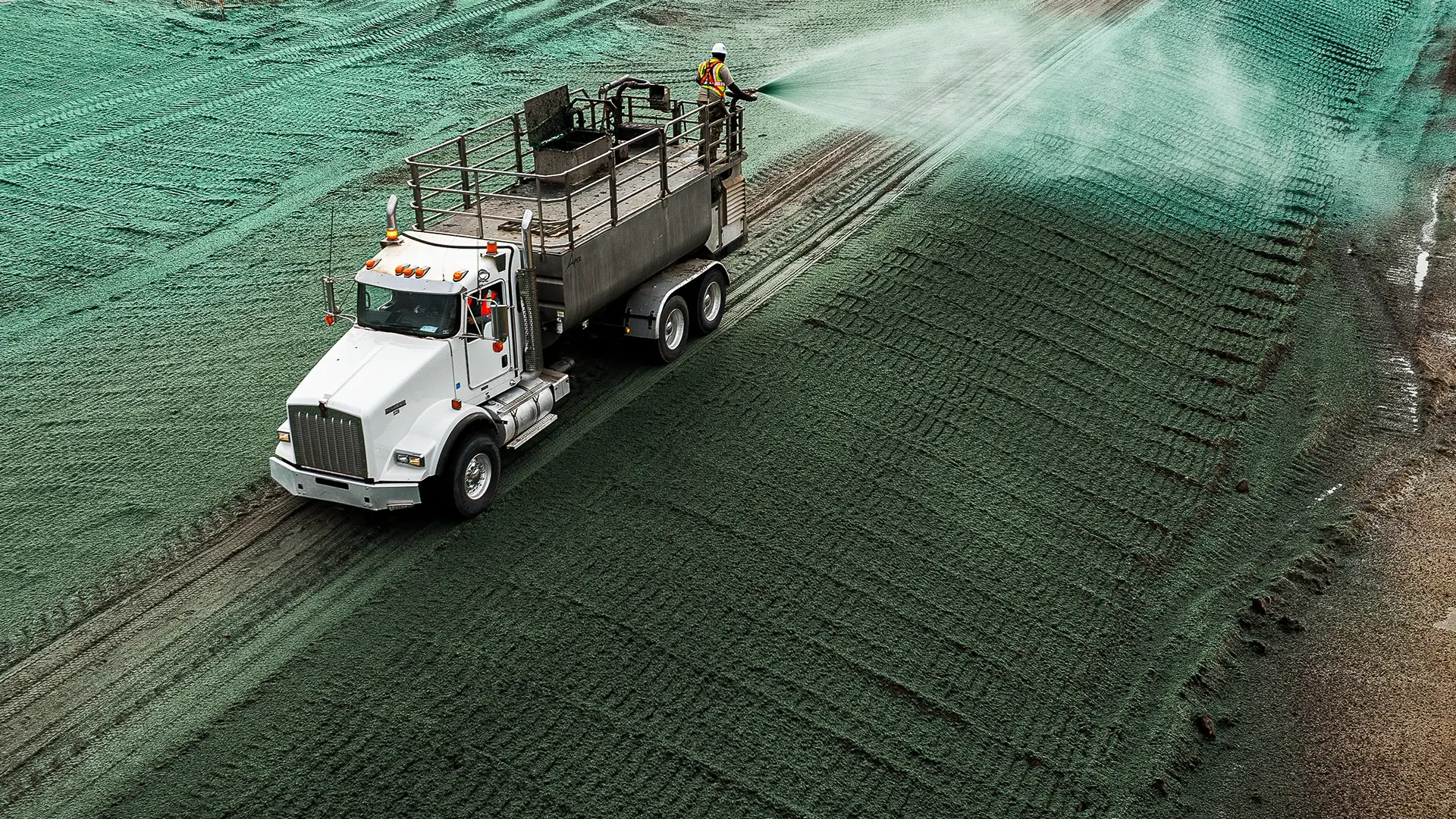A worker sprays green hydroseed from a large truck onto bare soil, likely for erosion control or grass seeding, covering the ground evenly on a construction or landscaping site.