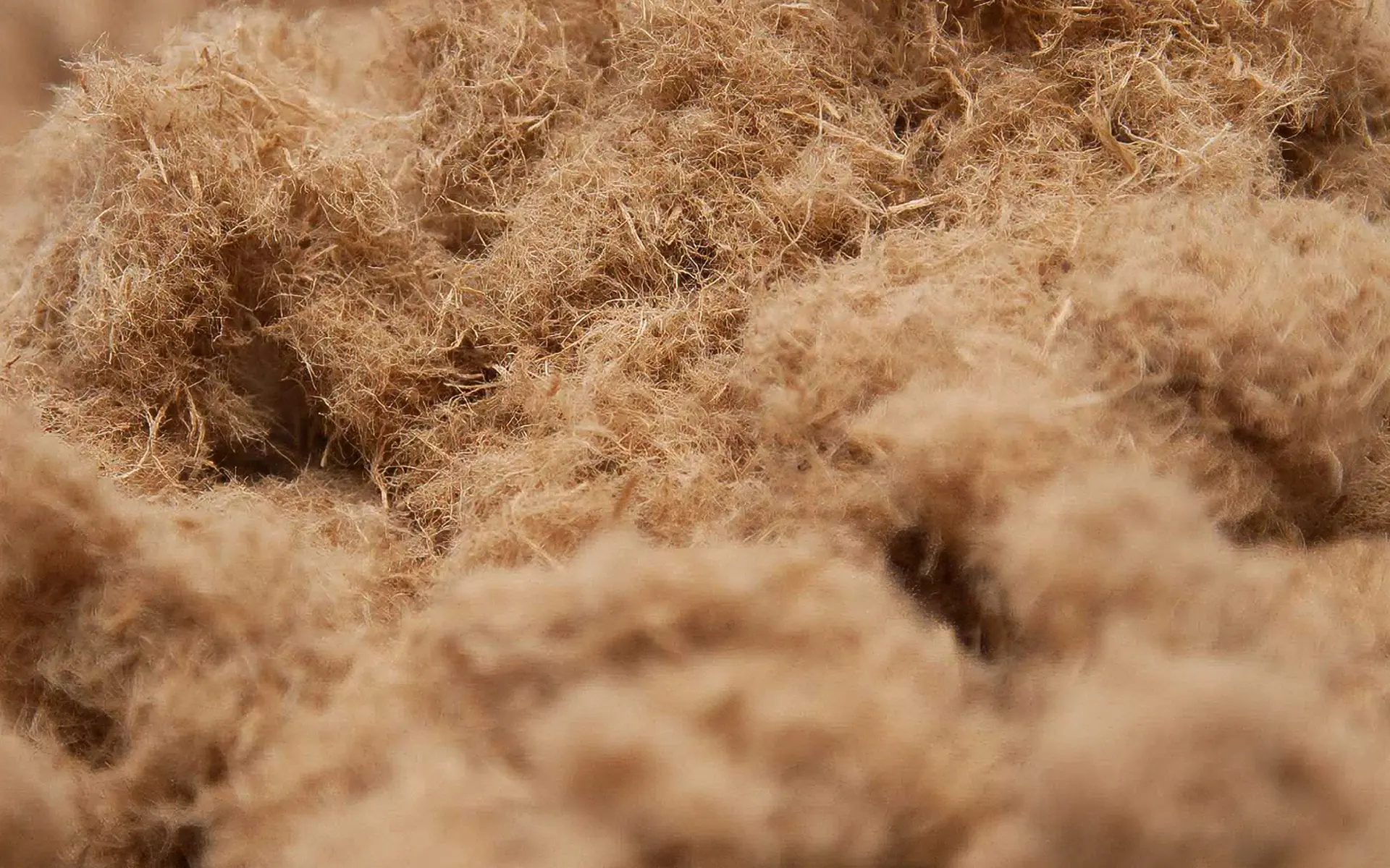 Close-up view of fluffy, light brown fibers clustered together, creating a soft, textured surface. The material appears natural and densely packed, with individual strands visible throughout the image.