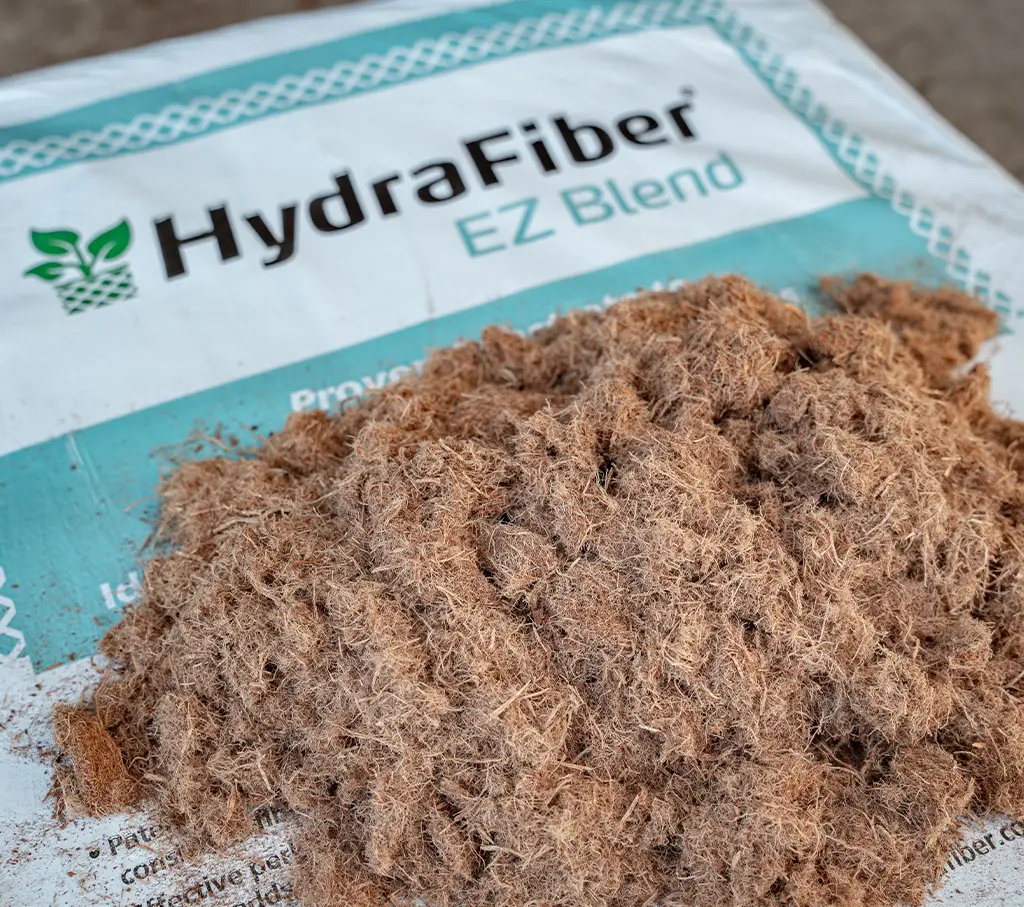 A pile of brown fibrous material sits on a white and blue HydraFiber EZ Blend bag, which has a green leaf logo and text on it.